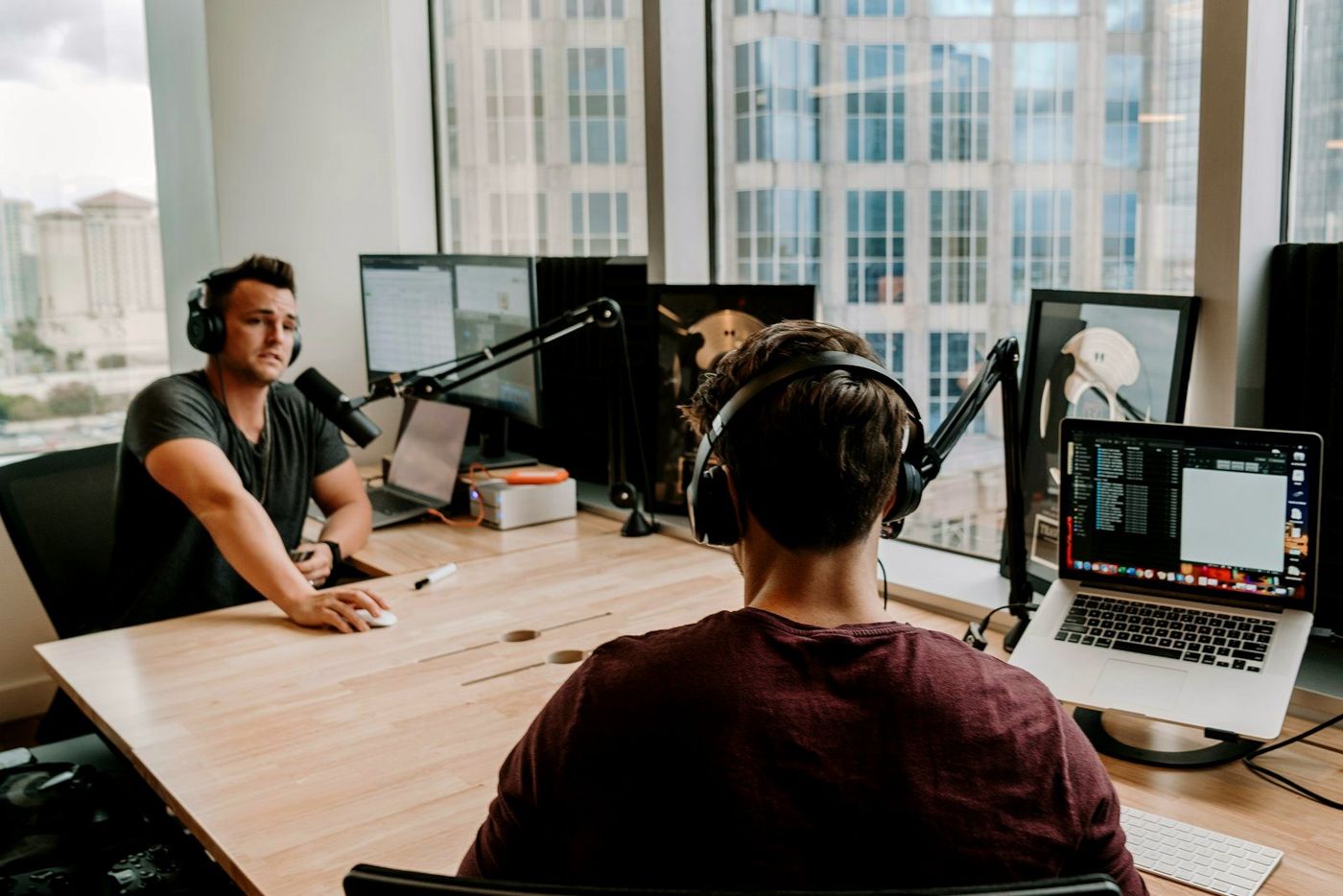 Two podcast hosts recording at a studio table with mics and laptops