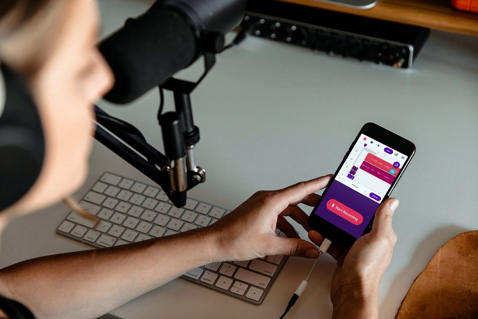 A podcaster at their desk scrolls messages on their phone, mic and keyboard beside them