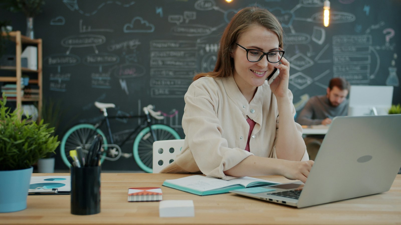 A woman on a phone call, smiling, at her laptop in a bright office