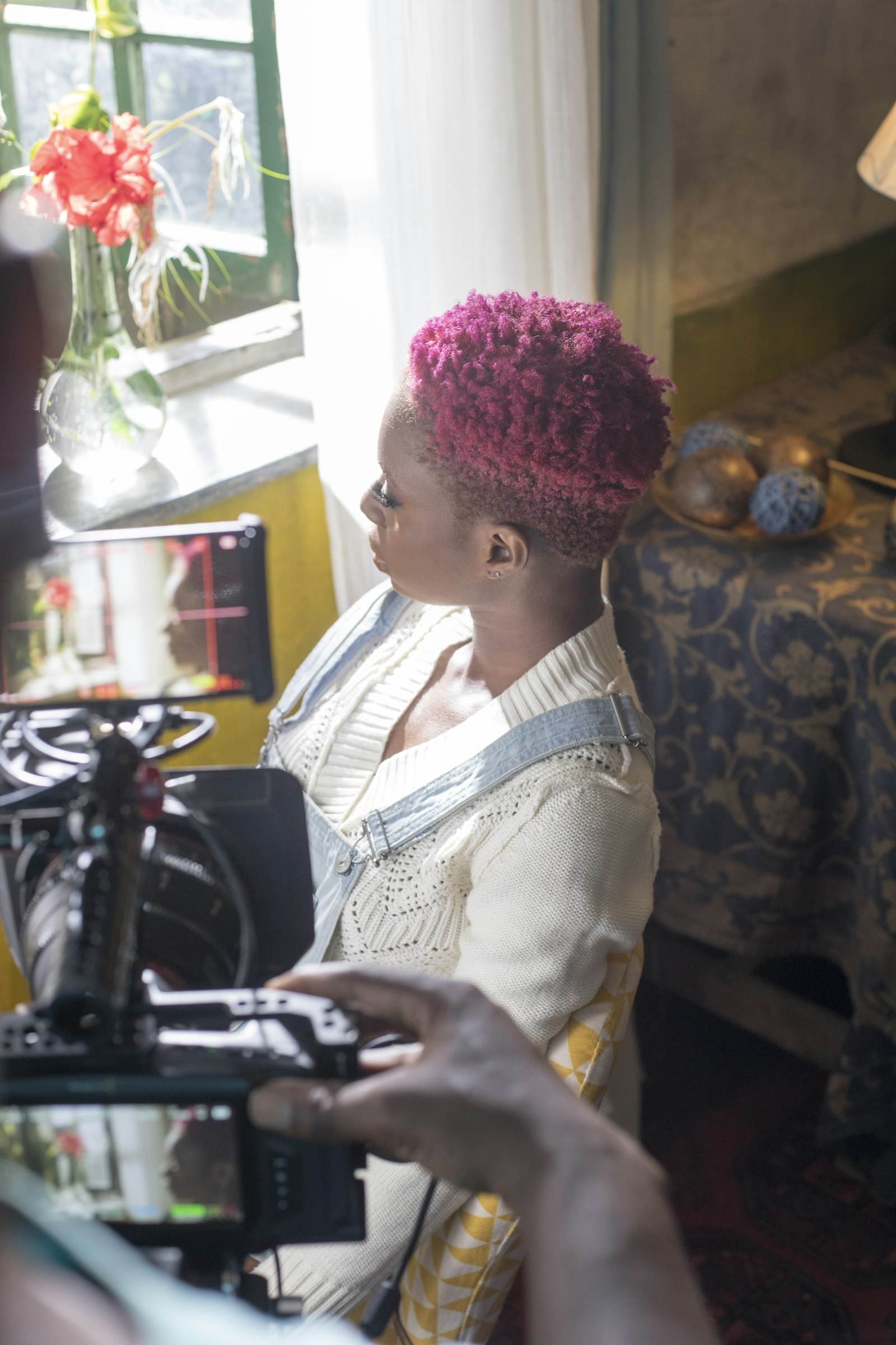A creator with short pink hair sits in front of a film camera, looking away from the lens.